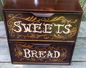 May include: A two-tiered wooden bread bin with a dark brown finish. The top tier is labelled "SWEETS" and the bottom tier is labelled "BREAD". Both tiers have gold painted floral designs.