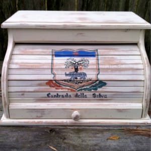 May include: White wooden bread box with a roll-top lid. The front of the box has a painted crest with a tree and the text "Contrada della Selva".
