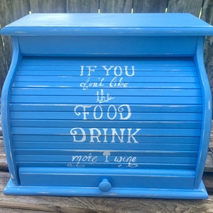 Op de afbeelding: Een blauwe houten broodtrommel met een roldeksel. Het deksel heeft een witte inscriptie met de tekst "Als je het eten niet lekker vindt, drink dan meer wijn".