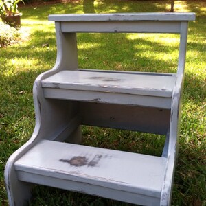 May include: A gray wooden two-step stool with a distressed finish. The stool has a top step and a bottom step. The stool is sitting on a grassy lawn.