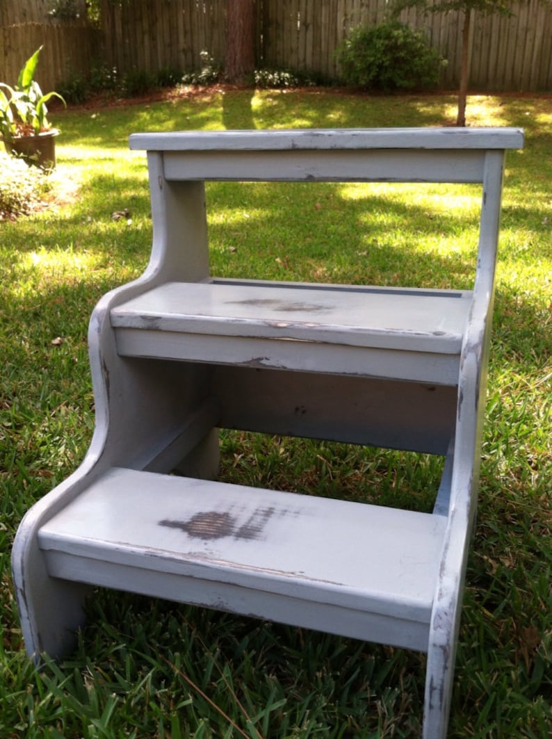 May include: A gray wooden two-step stool with a distressed finish. The stool has a top step and a bottom step. The stool is sitting on a grassy lawn.