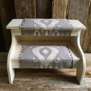 May include: White wooden step stool with two gray and white patterned fabric cushions. The stool has a simple design and is perfect for reaching high shelves.