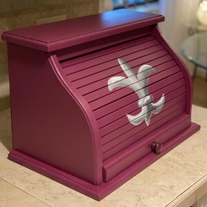 May include: A deep magenta bread box featuring a roll-top door with a hand-painted white and grey fleur-de-lis emblem. The wooden container has a flat top surface, a small round knob on the front panel, and a rectangular base with beveled edges.
