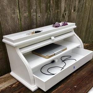 May include: White wooden shelf unit with a drawer, two shelves, and a top shelf. The unit has a curved front and a small knob on the drawer. There are sunglasses, keys, a tablet, and charging cords on the shelves.