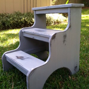 May include: A gray wooden two-step stool with distressed paint. The stool has a top step and a lower step. The stool is sitting on a grassy lawn.
