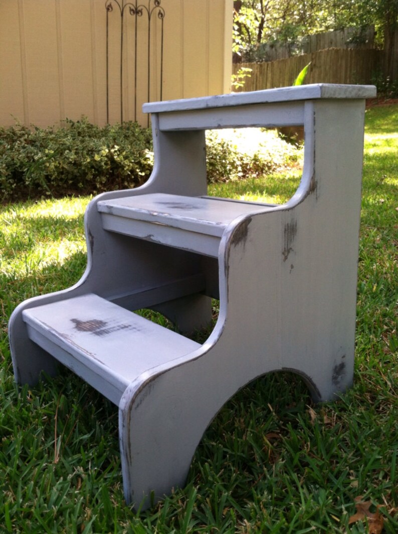 May include: A gray wooden two-step stool with distressed paint. The stool has a top step and a lower step. The stool is sitting on a grassy lawn.