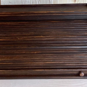 May include: A dark brown wooden bread box with a roll-top lid and a drawer. The box has a slightly curved top and a smooth finish.