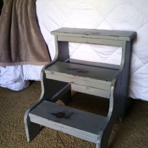 May include: A gray wooden step stool with two steps. The stool has a distressed finish and is slightly weathered.