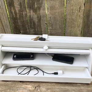 May include: White wooden charging station with two shelves and a drawer. The top shelf has a key and a key fob. The middle shelf has two cell phones. The bottom drawer has a charging cable and a plug.