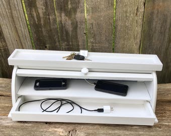May include: White wooden charging station with two shelves and a drawer. The top shelf has a key and a key fob. The middle shelf has two cell phones. The bottom drawer has a charging cable and a plug.
