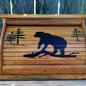 May include: A wooden bread box with a roll-top lid. The front of the box is painted with a black silhouette of a bear walking on a log in a forest setting.