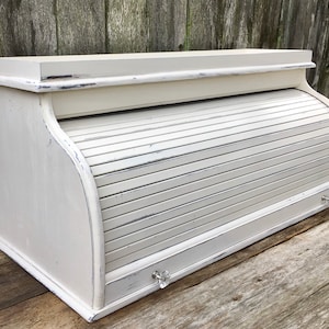 May include: White painted wooden bread box with a roll-top lid and two small knobs. The box has a distressed finish and is sitting on a wooden surface.
