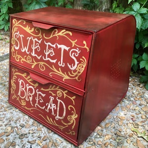 May include: A red metal bread box with two drawers. The top drawer is labeled "SWEETS" and the bottom drawer is labeled "BREAD". Both drawers have gold painted floral designs.
