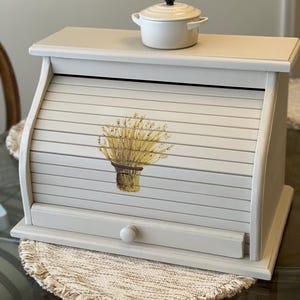 May include: A light beige wooden bread box with a roll-top door featuring a painted wheat sheaf design. A small, round, white ceramic lidded pot with a black knob sits on top of the bread box. The bread box has a single round knob on the front.