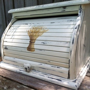 May include: White painted wooden bread box with a roll-top lid and a wheat design. The box has a distressed finish and a small knob on the front.