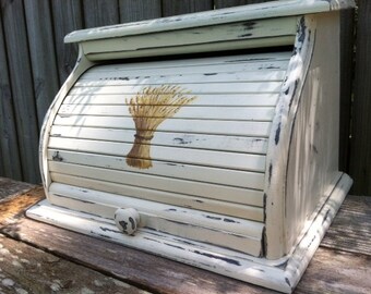 May include: White painted wooden bread box with a roll-top lid and a wheat design. The box has a distressed finish and a small knob on the front.