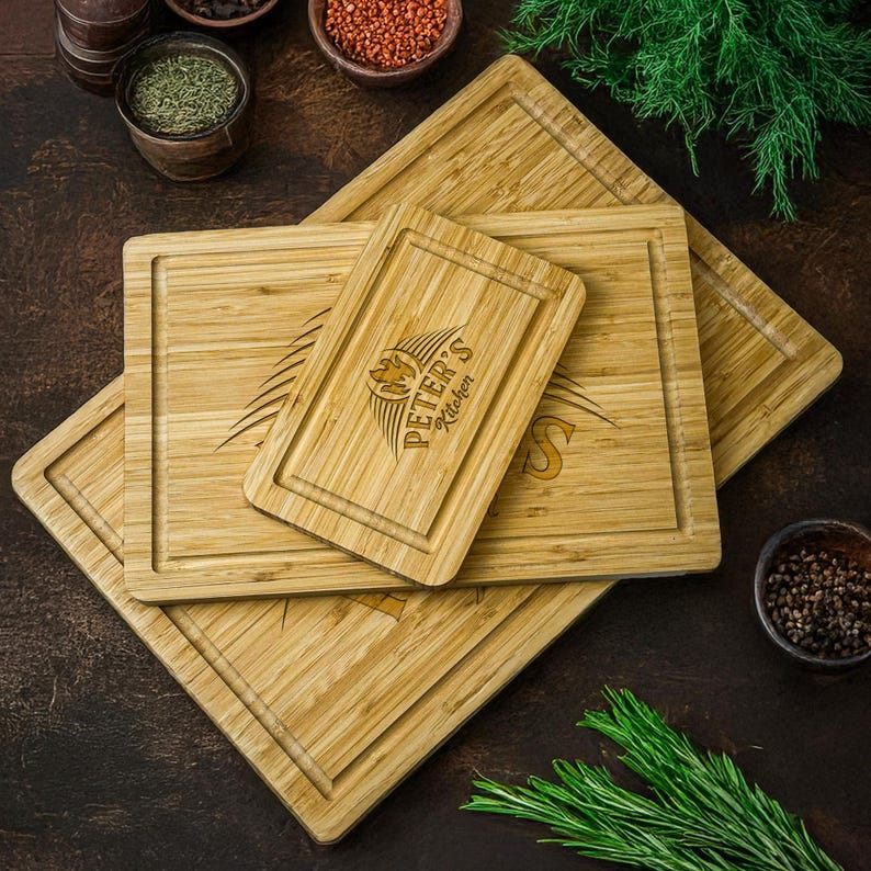 Może przedstawiać: Stos trzech bambusowych desek do krojenia. G&oacute;rna deska ma wygrawerowany napis "PETER'S Kitchen". Deski są jasnobrązowe, prostokątne i mają rowek na sok.