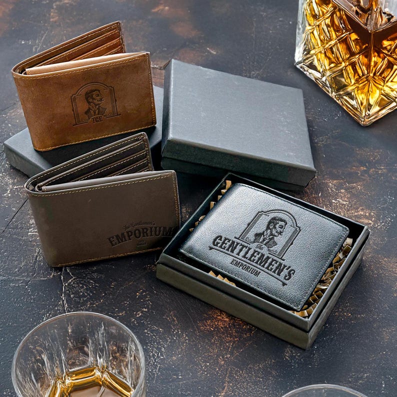 May include: Two brown leather wallets with engraved designs. One wallet has the text "The Gentleman's Emporium" and the other has the text "The Gentleman's Emporium".