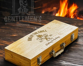 May include: A wooden box with a handle and metal clasps. The box is engraved with the text "King of the Grill" and a design of crossed grilling tools. The box is sitting on a wooden surface.
