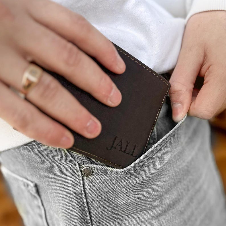 May include: A brown leather wallet with the name "JALL" embossed on the front. The wallet is being placed in a back pocket of gray jeans.