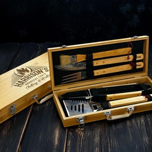 Op de afbeelding: Een bamboe barbecue gereedschapsset in een houten kist. De open kist toont roestvrijstalen en houten gereedschappen, zoals een spatel, vork, mes en borstel. De kist is voorzien van het opschrift "Harrison's Barbecue Tools Chilling & Grilling."