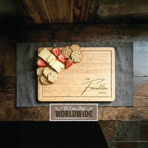 Könnte beinhalten: Ein rechteckiges Holzschneidebrett mit einer vertieften Kante, graviert mit "The Franklin Family". Das Brett ist mit Käse, Crackern und geschnittenen Erdbeeren belegt. Das Bild ist auf einem grauen Tuch platziert.