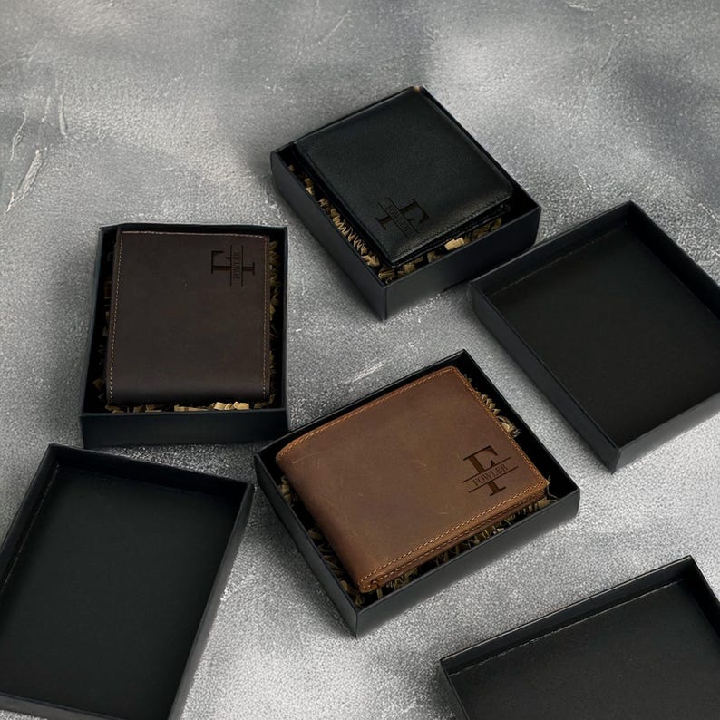 May include: Three leather wallets in black and brown with personalized initials in a black box. The wallets are displayed on a gray surface.