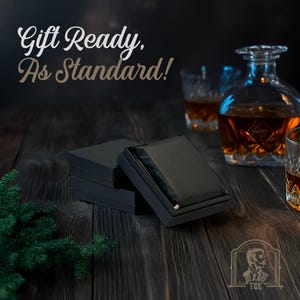 May include: A black gift box with a black ribbon and the text "Gift Ready, As Standard!" on a dark wood surface. There are two glasses of amber liquid and a decanter of amber liquid in the background.