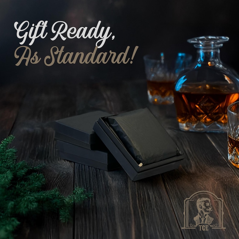 May include: A black gift box with a black ribbon and the text "Gift Ready, As Standard!" on a dark wood surface. There are two glasses of amber liquid and a decanter of amber liquid in the background.