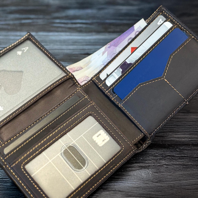 May include: A brown leather wallet with a stitched design. The wallet is open and has several card slots, a billfold, and a clear ID window. The wallet is filled with cards and cash.