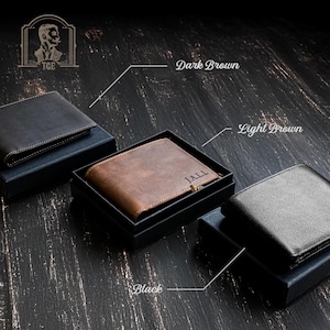 May include: Three leather wallets in different colors: black, dark brown, and light brown. The wallets are displayed on a wooden surface. The light brown wallet has the name "JALL" engraved on it.