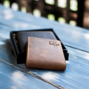 May include: Two leather wallets, one brown and one black, are displayed on a light blue wooden surface. The brown wallet has a small, embossed logo on the front.