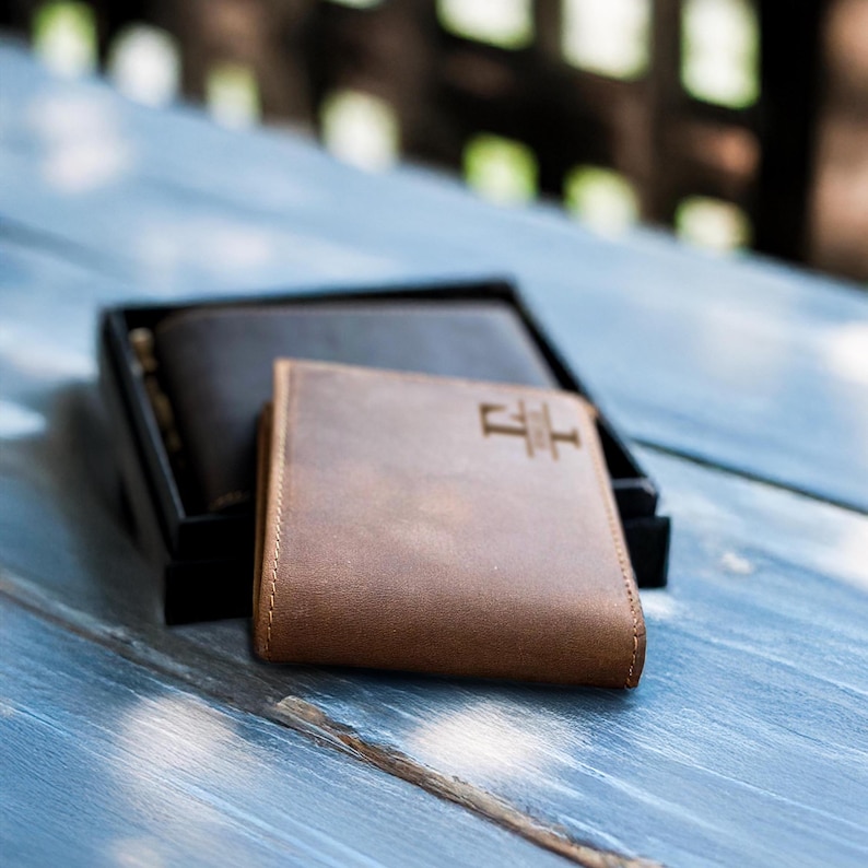 May include: Two leather wallets, one brown and one black, are displayed on a light blue wooden surface. The brown wallet has a small, embossed logo on the front.