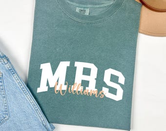 May include: A blue spruce t-shirt with the word "MRS" in large white block letters and the name "Williams" in a cursive font. The shirt is folded, with a pair of blue jeans and a tan hat in the background.