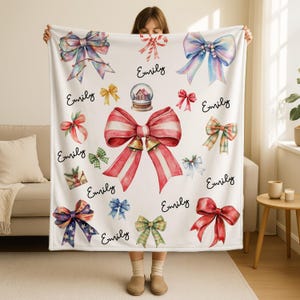 May include: A white blanket featuring watercolor illustrations of various bows in red, blue, green, and yellow. The word "Emily" is written in cursive script throughout the design. A snow globe is also featured.