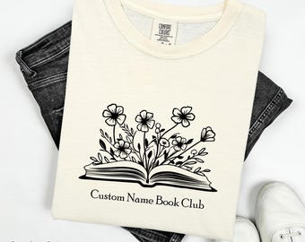 Peut inclure: T-shirt ivoire Comfort Colors avec un motif de livre floral noir. Le graphisme du livre ouvert présente des fleurs et le texte "Custom Name Book Club". Le t-shirt est posé sur un jean en denim foncé et des baskets blanches.