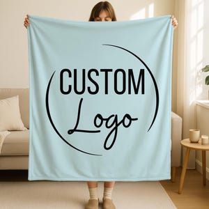 May include: A light blue blanket with the words "CUSTOM Logo" in black, inside a black semi-circle. The blanket is held up by a person. The blanket is a solid color, with a soft, plush appearance.