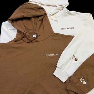 May include: Two hooded sweatshirts, one brown and one cream. The brown sweatshirt has the Roman numeral 'XXXVII MMXXIII' embroidered on the front. The cream sweatshirt has the same Roman numeral on the front and the letters 'T' and 'E' with hearts on the sleeves.