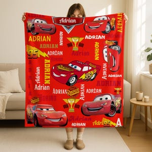 May include: A red blanket featuring the character Lightning McQueen from the movie Cars. The blanket has the name "Adrian" printed in white, along with images of the race car and the number 95. The blanket is a vibrant red color.