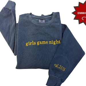 Pode incluir: Um moletom azul com as palavras "girls game night" bordadas em amarelo no peito. A manga tem "est. 2018" bordado em amarelo. Um gr&aacute;fico vermelho em forma de estrela com a palavra "EMBROIDERED" est&aacute; no canto superior direito.