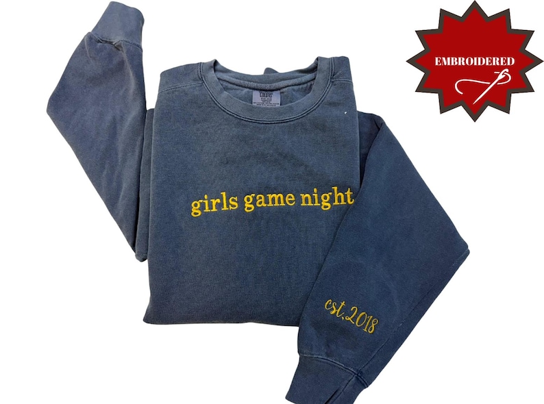 Pode incluir: Um moletom azul com as palavras "girls game night" bordadas em amarelo no peito. A manga tem "est. 2018" bordado em amarelo. Um gr&aacute;fico vermelho em forma de estrela com a palavra "EMBROIDERED" est&aacute; no canto superior direito.