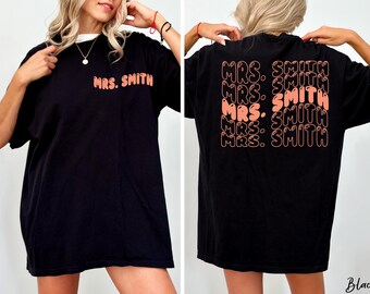 May include: Black oversized t-shirt with the text "MRS. SMITH" in a retro orange font on the front and back. The back of the shirt features the text repeated multiple times. The shirt has a white collar.