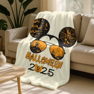 May include: Cream-colored Halloween-themed throw blanket. The design features black Mickey Mouse ears with orange bats and a spiderweb. Sunglasses with orange lenses show a haunted house scene. The text "HALLOWEEN 2025" is printed below.
