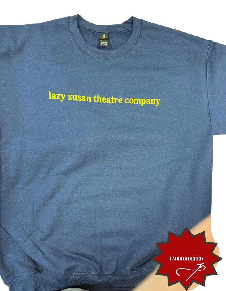 Pode incluir: Camisola azul marinho com gola redonda e o texto amarelo "lazy susan theatre company". A camisola tem um design cl&aacute;ssico e &eacute; feita de um material macio. Uma etiqueta bordada vermelha tamb&eacute;m &eacute; vis&iacute;vel.