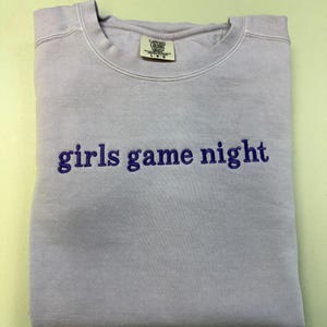 Pode incluir: Uma camisola dobrada, de cor lil&aacute;s claro, com o texto bordado "girls game night" em roxo mais escuro. A camisola tem gola redonda e uma etiqueta na gola.