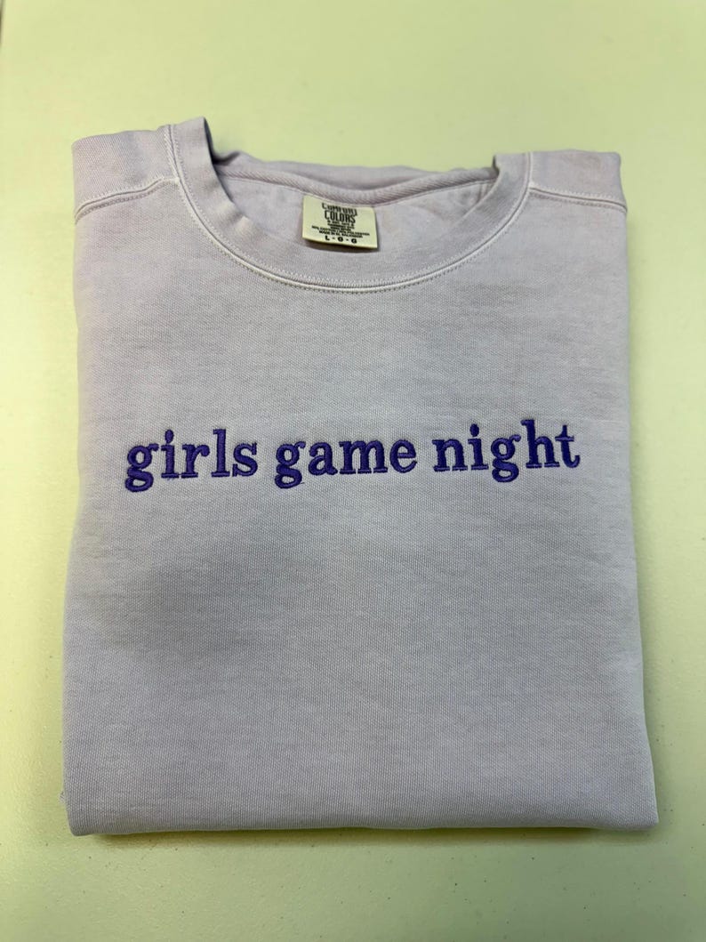 Pode incluir: Uma camisola dobrada, de cor lil&aacute;s claro, com o texto bordado "girls game night" em roxo mais escuro. A camisola tem gola redonda e uma etiqueta na gola.