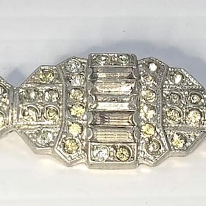 May include: A silver-toned vintage brooch with a geometric design. The brooch features a central rectangular section flanked by decorative, fan-shaped ends. It is embellished with clear and yellow rhinestones.