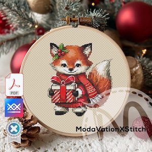 Puede incluir: Bordado de punto de cruz de un zorro con una bufanda roja y una caja de regalo. El zorro tiene pelaje naranja, orejas negras y una cola con punta blanca. El diseño está en un bastidor de bordado de madera. El fondo incluye adornos navideños y vegetación.