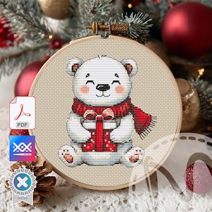 May include: Cross-stitch embroidery of a smiling polar bear wearing a red scarf and holding a red gift box. The design is set in a wooden embroidery hoop. The background includes Christmas ornaments and a pine cone.