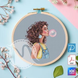 May include: Cross-stitch embroidery in a wooden hoop features a woman blowing a bubble. The woman has brown curly hair, blue earrings, and is wearing a yellow top and blue overalls. The bubble is pink and translucent. The background is light blue.